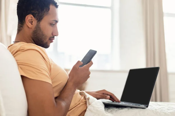 Side View Of Middle Eastern Man Using Phone And Laptop Computer Communicating And Working Online At Home. Technology And Internet, Freelance Career Concept