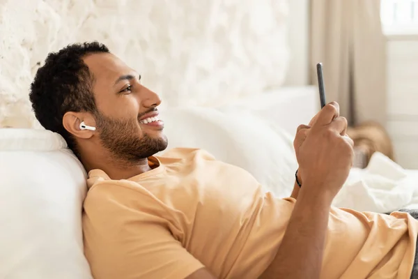 Cheerful Arabic Man Listening To Music Using Smartphone And Wearing Earbuds Lying In Bed In Modern Bedroom At Home. Guy Using Musical Application Relaxing In The Morning. Side View