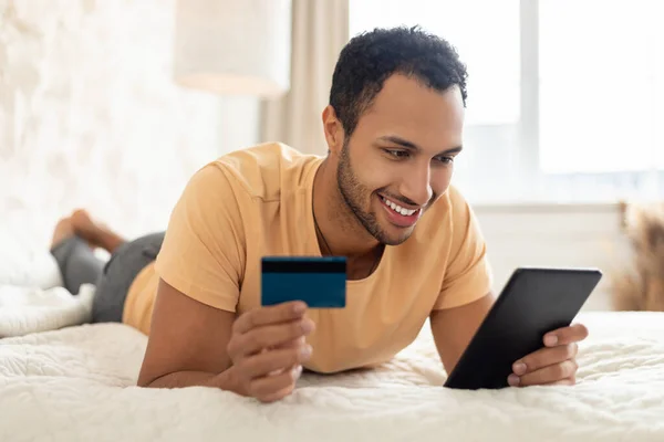 Contented Middle Eastern Male Buyer Shopping Online Using Digital Tablet And Credit Card Lying On Bed In Modern Bedroom Indoors. Guy Making Payment Via Internet Banking At Home. Ecommerce Concept