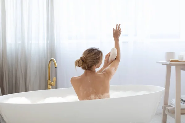 自宅で泡風呂に入る魅力的なブロンドの大人の女性のバックビュー スポンジで彼女の美しい腕をこすり 週末を楽しんで 彼女の肌の世話をし スペースをコピーします ボディケアの概念 — ストック写真