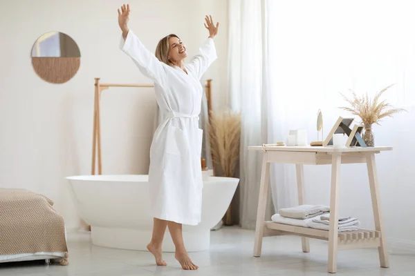 Joyful attractive middle aged lady with blonde hair dancing with hands up by white bathroom at home, wearing long bathrobe, enjoying weekend, going to take relaxing bath, panorama, copy space