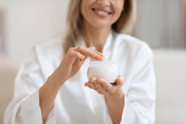 Cropped of blonde woman in white silky bathrobe holding white jar with cream, unrecognizable lady using face cream or body lotion after shower, skin care at home concept