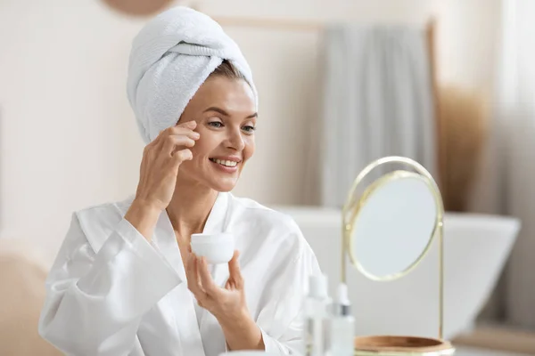 シルクバスローブとターバンで美しい大人の女性は シャワー後の栄養アイクリームを使用して 鏡と笑顔を見て 顔のケアルーチンを楽しんで コピースペース 暗い円 目のしわ治療 — ストック写真