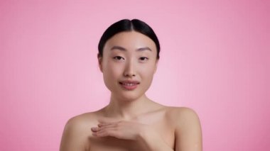 Genç çekici, bakımlı Asyalı kadın yakasına dokunuyor, cildinin keyfini çıkarıyor, pembe stüdyo arka planı, yavaş çekim.
