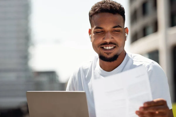 Happy black man working with documents and laptop computer outdoors, sitting in city area, using pc and preparing report. African american guy studying outside