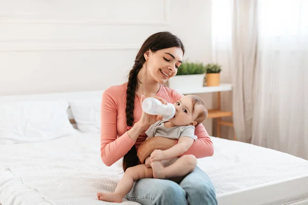 Baby care. Caring mother giving bottle with milk to her infant child while sitting on bed at home, young mom enjoying spending time with her kid