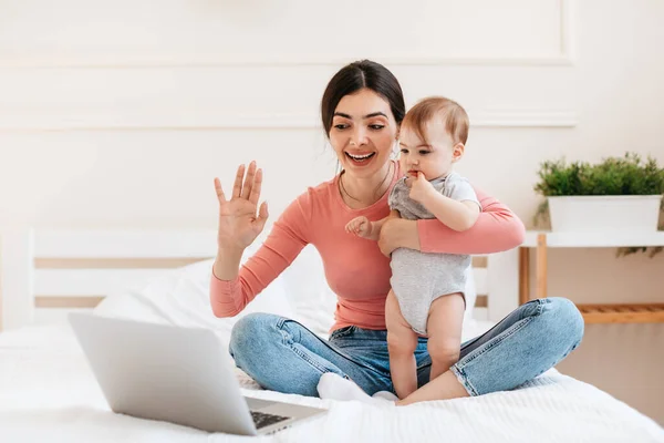 Happy mom holding baby and making video call with laptop, sitting on bed at home and waving hand at webcamera. Happy mother with infant child communicating with family online