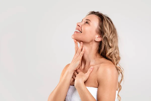 Double Chin Treatment. Beautiful Middle Aged Woman Touching Soft Smooth Skin On Neck, Attractive Mature Lady Standing Wrapped In Towel Over Light Grey Background, Enjoying Her Beauty, Copy Space