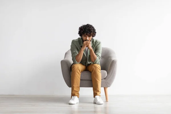 Depressed curly bearded young indian guy in casual outfit sitting in armchair over white studio background, looking down, thinking about something, suffering from loneliness, copy space, full length