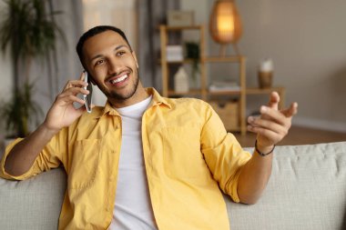 Afro-Amerikalı olumlu genç adam akıllı telefonda konuşuyor, evdeki kanepede oturuyor, boş yer var. Milenyum siyahi adam telefonla konuşuyor. Oturma odasında dostça sohbet ediyor.
