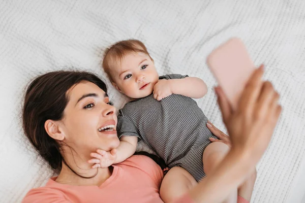 어머니가 귀여운 아기와 셀카를 데려가 침대에 아기와 잠자리에서 핸드폰으로 사진을 — 스톡 사진