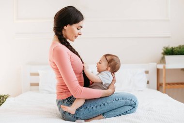 Küçük çocukla oynayan genç bir anne, annesinin kucağında oturan sevimli bir çocuk ve saçlarına dokunuyor. Çocuk bakımı ve anne olmanın neşesi