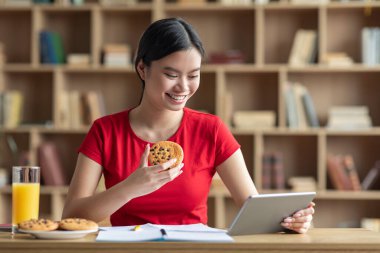 Gülümseyen genç Çinli kadın kurabiye yiyor ve içerideki masadaki tablete bakıyor. Sosyal ağlar, evde eğitim, blog, modern cihazlar, covid-19 salgını sırasında internet üzerinden reklam ve teklif