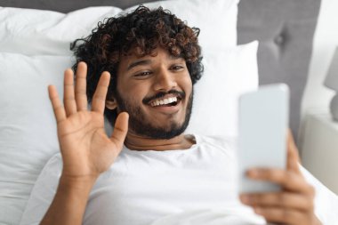 Sevgilisiyle video görüşmesi yapan yakışıklı doğulu adam, yatakta uzanıyor, cep telefonunu tutuyor ve kamera önünde el sallıyor, yakın plan fotoğraf çekiyor. Haberleşme ve modern teknolojiler