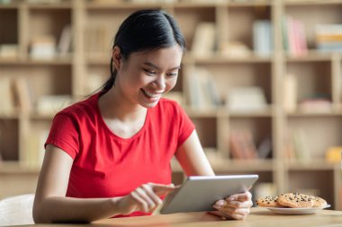 Sınıfta ya da kütüphanenin içinde tablet yazan zeki Koreli kız öğrenci gülümsemesi. Evde eğitim, sosyal medya sohbeti uygulaması, blog, harika bir teklif ve covid-19 salgını sırasında aletle çalışma