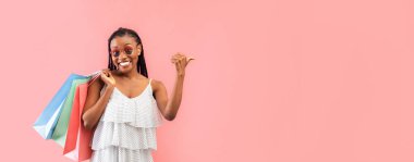 Alışveriş torbaları ile neşeli siyah kadın gülümseyen ve boş alanı işaret eden pembe stüdyo arka planı, pankart tasarımı. Afro-amerikalı bayanın elinde alışveriş yeri ve reklam yeri var.