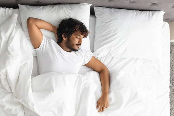 Top view of handsome bearded young indian guy in pajamas resting at home, sleeping on his back with arm behind head under blanket in comfortable bed., copy space Healthy sleeping concept