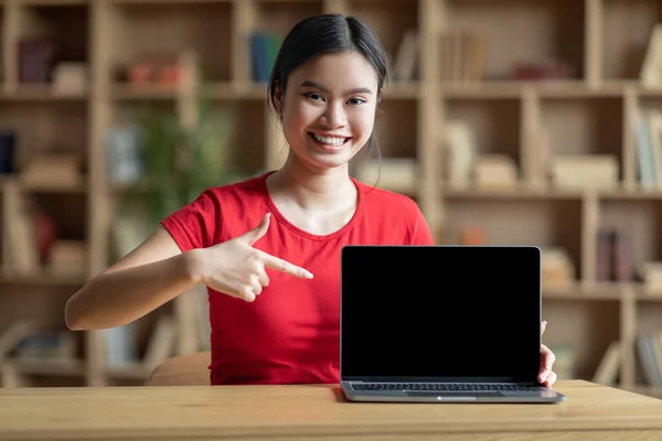 Smiling teen korean lady point finger at computer with blank screen for study in room interior. Modern education at home, work remote, device for online lesson and good website due covid-19 pandemic