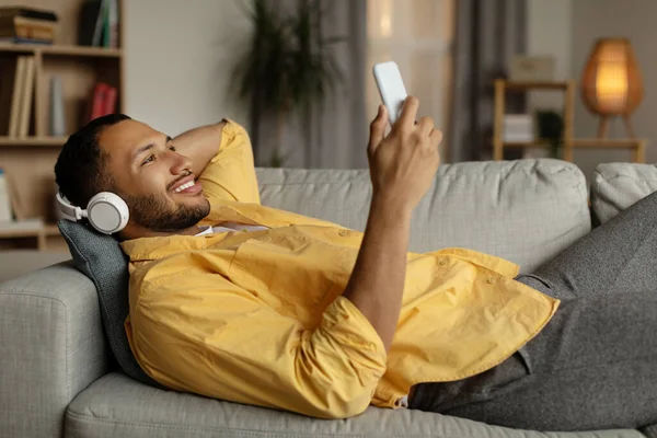 自宅でソファに横たわって スマートフォン上でオンライン音楽を聞いて無線ヘッドフォンでハンサムな若い黒人男性 幸せなアフリカ系アメリカ人男性でヘッドセットビデオを見ます上の携帯電話 — ストック写真