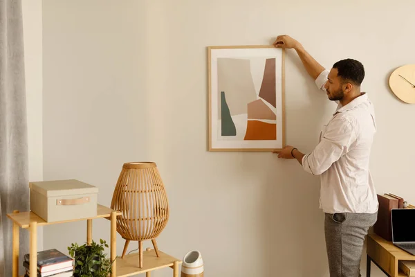 Modern home decor and interior design. Young black man hanging painting, putting picture frame on wall indoors, copy space. Millennial African American guy decorating his apartment