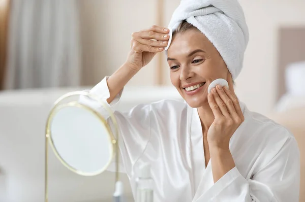 Good-looking adult lady in white bathrobe and turban on her head looking at mirror and using peeling pads, enjoying beauty routine at home, closeup photo, copy space. Face peeling concept