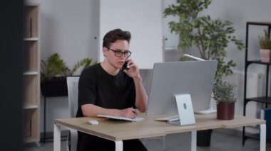 Mesafeli iş kavramı. Genç, zeki adam yönetici gözlük takıyor, müşteriye danışıyor ve not defterine notlar yazıyor, modern iş yerinde oturuyor, ağır çekimde.