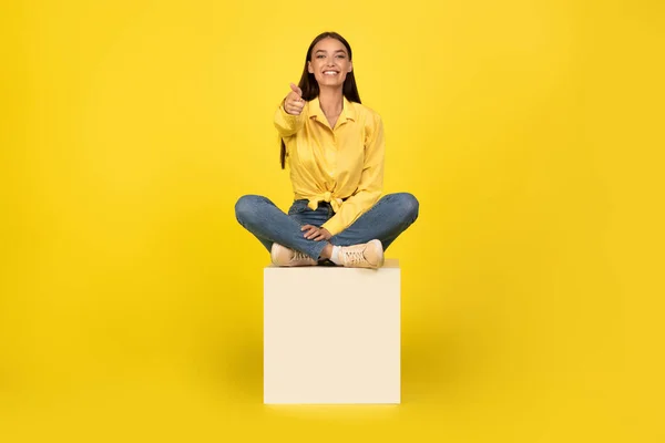 快乐的年轻女士指出手指微笑相机选择你坐在立方体上而不是黄色背景 广告概念 全长铅球 — 图库照片
