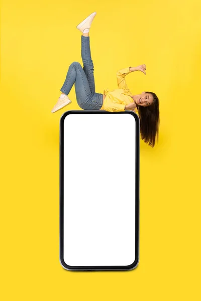 巨大な携帯電話の上に横たわる陽気な女性は 黄色のスタジオの背景に空白のスマートフォン画面広告モバイルアプリケーションで親指を指しています 縦型モックアップ — ストック写真