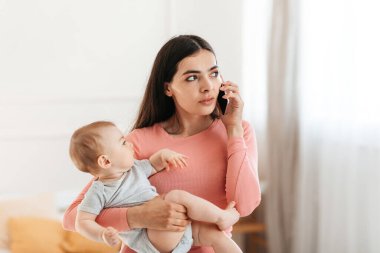 Genç annelerin yaşam tarzı. Düşünceli bir anne cep telefonuyla konuşuyor ve kucağında bebek, doktoru arıyor ya da evde telefon görüşmesi yapıyor.