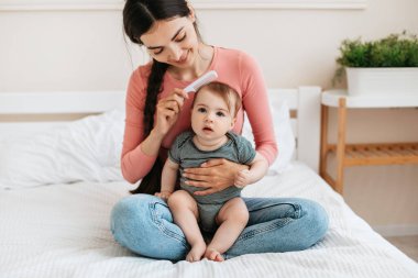 Annenin saçını taraması, çocuğun annesinin kucağına oturması ve kameraya bakması annelik iznini ve çocuk bakımını seven bir kadın. Sevimli anne ve çocuğu evde kaynaşıyor.