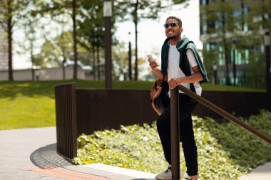 Güneşli bir günde cep telefonuyla gezen, sırt çantası takan, güneş gözlüğü takan, boş zamanların tadını çıkaran ve gülümseyen, uzay kopyası olan şık siyahi adam.