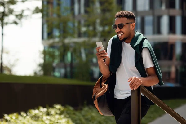 On my way to work. Happy stylish african american man using his smartphone and walking in park area, texting on phone and smiling, panorama with free space