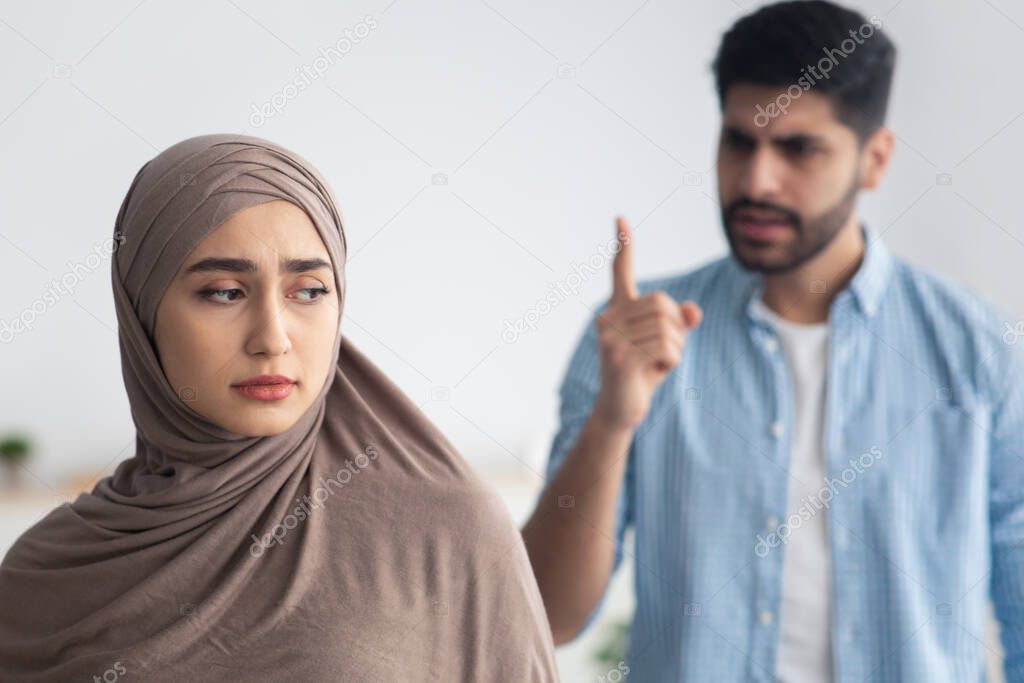 Angry Husband Shouting At Unhappy Muslim Wife Standing At Home. Selective Focus On Sad Woman In Hijab Suffering From Domestic Abuse And Violence. Bad Marriage, Unhappiness Concept