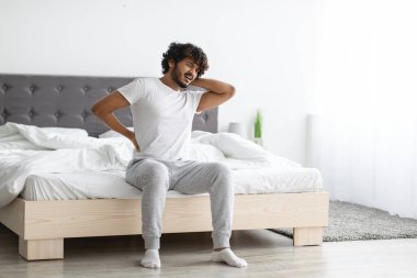 Yatak odasında sırt ağrısı çeken mutsuz, genç Hintli adam, sabah uyandığında, boynuna ve beline iki eliyle dokunuyor, pijama giyiyor, ev içi, fotokopi odası