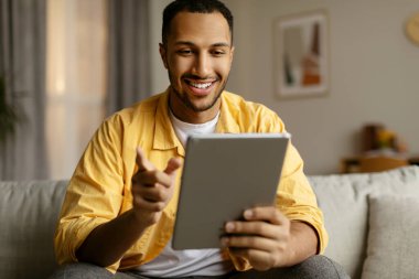 Tabletli mutlu genç siyahi adam kanepede oturuyor, çalışıyor ya da internette çalışıyor, evde görüntülü sohbet ediyor. Neşeli Afrikalı Amerikalı adam internetten iletişim kuruyor.