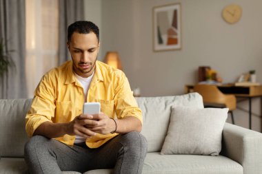 Akıllı telefon uygulaması kullanan, evdeki kanepede oturan, boş alan kullanan ciddi bir zenci. Milenyum Afrikalı Amerikalı adam cep telefonuyla interneti tarıyor. Gadget yaşam tarzı, mobil uygulamalar. Boşluğu kopyala