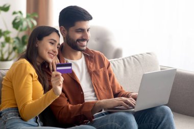 İnternetten alışveriş yapan mutlu Arap Alıcılar dizüstü bilgisayar ve kredi kartı ile ödemeyi evde yapmayı kucaklıyor. Orta Doğulu eşlerin bir şey satın almasının yan görüntüsü. Ticaret için. Yan Görünüm