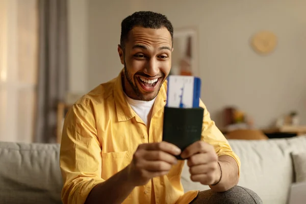 Excited young black man with passport and tickets getting ready for summer vacation, sitting on couch at home. Cheerful African American guy feeling happy about tourist trip indoors