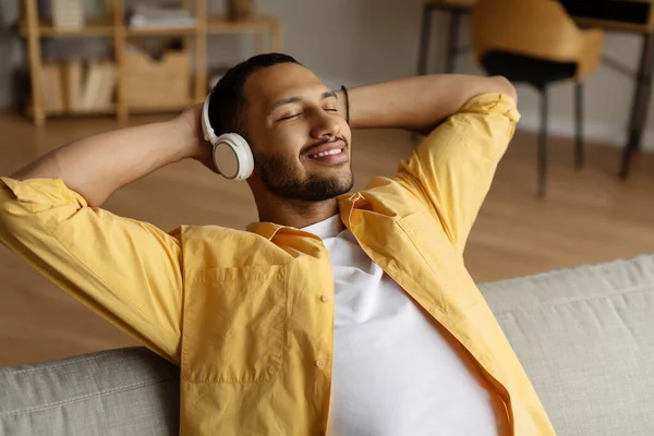 ヘッドフォンで陽気な若い黒人男性のソファの上に目を閉じて 自宅で音楽を聞いて座っている 千年のアフリカ系アメリカ人の男は ヘッドセットで新しいオーディオブックを楽しんで 屋内でリラックス — ストック写真