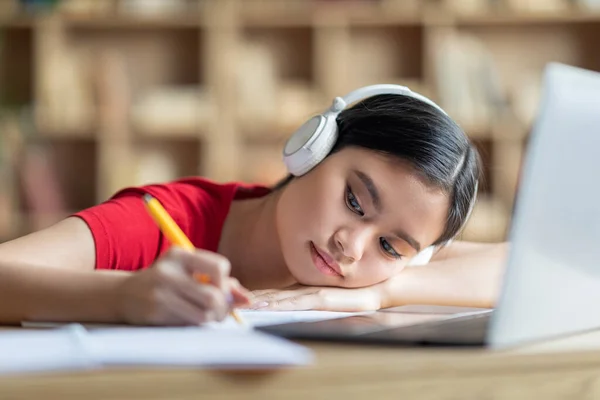 疲惫的日本少女戴着耳机做笔记 靠着笔记本电脑躺在桌旁 在房间里学习 在Covid 19大流行病期间 工作负担过重 教育问题 在家远程录像教学 — 图库照片