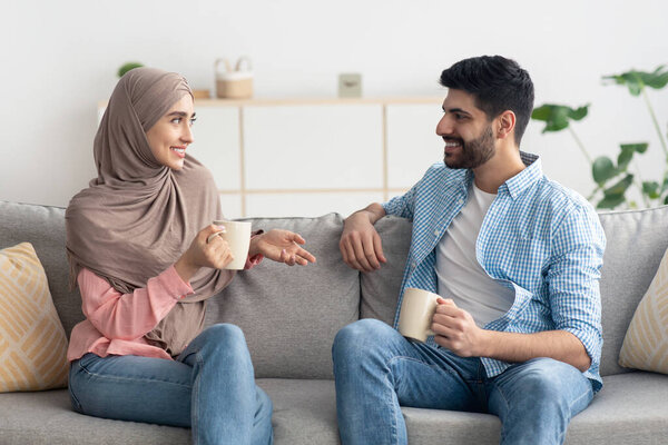 Cheerful Middle Eastern Spouses Drinking Coffee And Talking Flirting Sitting On Sofa At Home. Muslim Husband And Wife Enjoying Conversation Holding Mugs Indoor. Marriage And Relatioship Concept