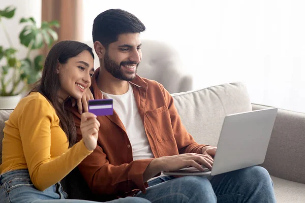 Happy Arabic Buyers Couple Shopping Online Via Laptop And Credit Card Embracing Making Payment Sitting At Home. Side View Of Middle Eastern Spouses Purchasing Something. Ecommerce. Side View