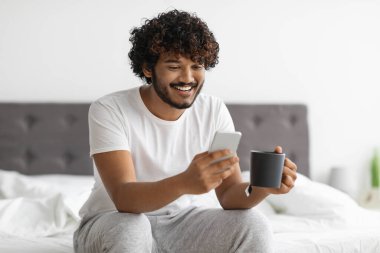 Evde pijama giymiş, kahve içip akıllı telefon kullanan hafta sonları tek başına takılıp, sosyal medyayı kontrol eden mutlu ve yakışıklı bir doğulu adam. Boş zaman ve aletler.