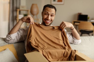 Genç siyahi adam karton kutuyu açıyor, iyi teslimat servisinden memnun, online mağaza siparişi alıyor, evdeki kanepede oturuyor. Milenyum Afrikalı Amerikalı adam oturma odasında paketleri açıyor.