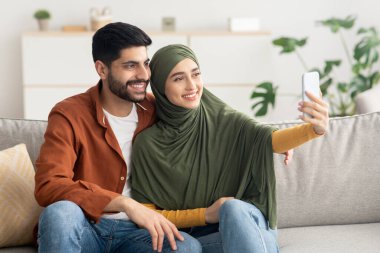 Mutlu Müslüman koca ve eş akıllı telefonda selfie çekiyor koltukta oturuyor, poz veriyor ve evde eğleniyor. Teknoloji ve Gadget, Aile Yaşam Tarzı Konsepti