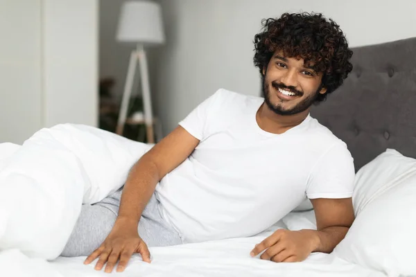 穿着睡衣的快乐的热胡子卷曲印地安人独自躺在床上 摸着身边空旷的空间 对着相机和卧室的内部微笑 千禧年中的性健康概念 — 图库照片