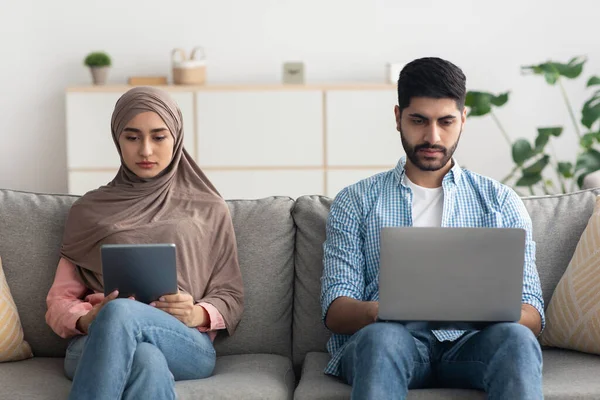Islamic Husband And Wife Using Digital Tablet And Laptop Sitting Together And Websurfing On Weekend At Home. Middle Eastern Couple Using Computers Posing On Sofa Indoor. Technology And Internet