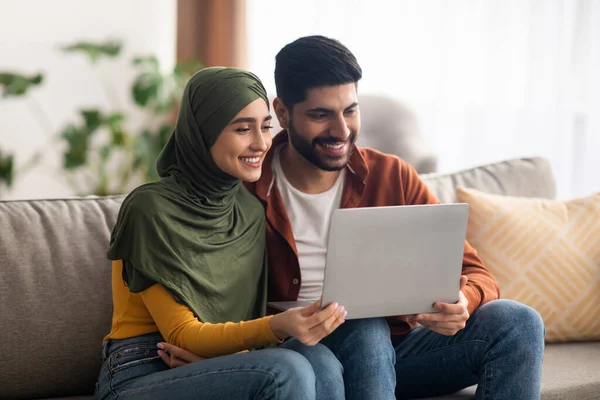 Middle Eastern Couple Using Laptop Sitting In Modern Living Room At Home. Happy Arabic Spouses Browsing Internet Together Watching Film Online Posing On Sofa Indoor. Internet And Technology