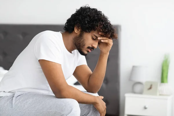 Depressed curly bearded young indian guy in pajamas sitting on bed alone at home, touching his head, thinking about something, suffering from loneliness, side view, copy space