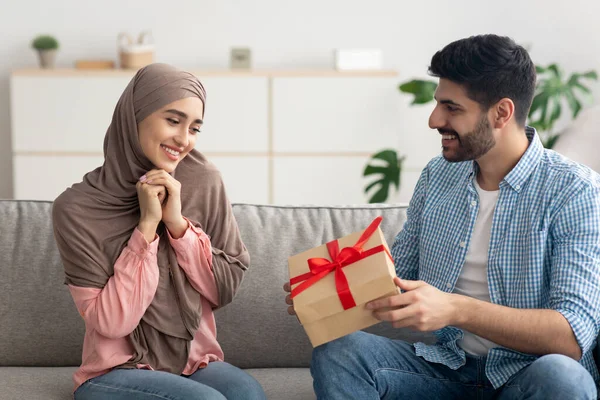 Family Holiday. Muslim Husband Giving Gift To Wife Celebrating Her Birthday At Home. Happy Middle Eastern Woman Receiving Wrapped Present Box From Man Sitting On Sofa Indoor